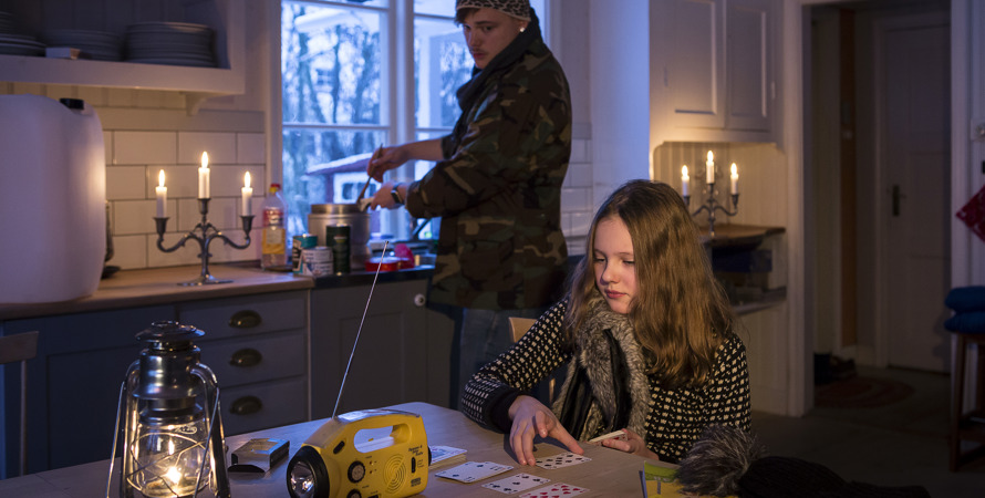 Ett barn och en vuxen är påklädda inne. Elen fungerar inte, det står en lykta och en ficklampa på bordet.