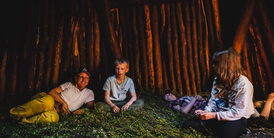 En lärare halvligger på björkris i en kåta. Två barn sitter och ett ligger ner.